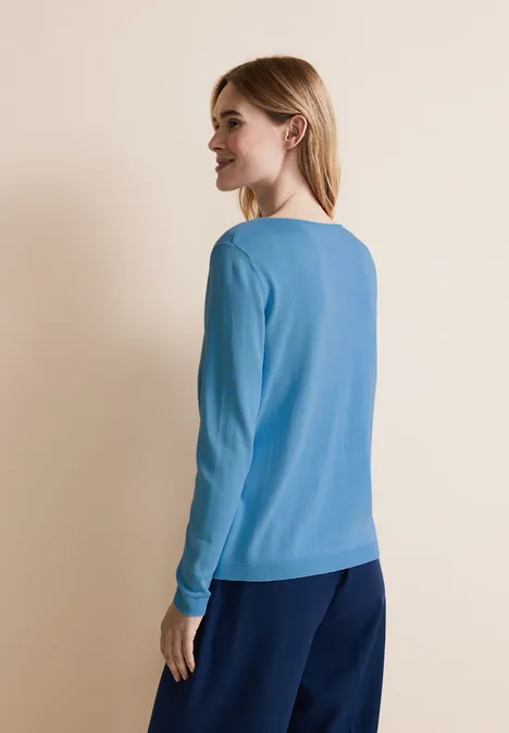 Frau mit blondem Haar trägt blauen Pullover, steht seitlich vor beigem Hintergrund.